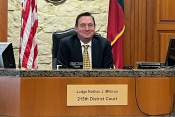 Un hombre sonriente con traje y corbata, identificado como el Juez Nathan J. Milliron, sentado en un estrado de tribunal con banderas detrás