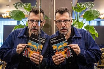 Hombre con gafas y barba, chaqueta de mezclilla, sostiene un libro. Se observa su reflejo en un espejo. Fondo con plantas y mobiliario
