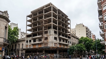 Edificio de hormigón abandonado de siete pisos en una calle concurrida, rodeado de otros edificios y árboles, con personas caminando en la acera.