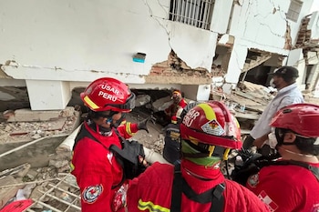 Cuatro bomberos del equipo USAR Perú con uniformes rojos y cascos frente a un edificio blanco parcialmente colapsado y escombros en el suelo