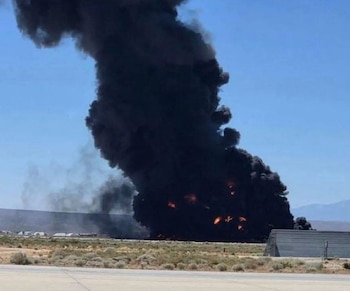 Gran columna de humo negro espeso y llamas anaranjadas se eleva hacia un cielo azul claro desde una explosión en un paisaje árido