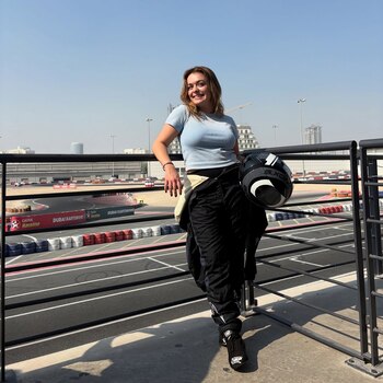 Izzy Hammond sonríe, vestida con un traje de carreras negro y una camiseta azul, sosteniendo un casco, con la pista de karts de Dubái de fondo