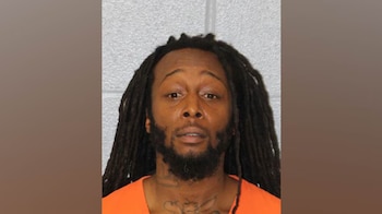 Foto policial de un hombre afroamericano con dreadlocks y barba, mirando al frente, vistiendo una prenda de color naranja, sobre un fondo gris claro