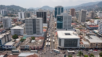 Vista aérea de una ciudad con muchos edificios de diferentes alturas, una calle central con abundante tráfico de vehículos y personas, y montañas al fondo.