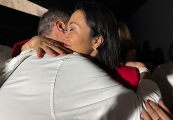 Dos personas se abrazan, una mujer de cabello oscuro con camisa roja y arete de perla, y un hombre con camisa blanca y pulsera. Uñas rojas de la mujer