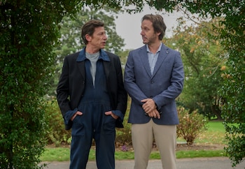 Dos hombres parados, uno con blazer azul y pantalones beige, el otro con blazer negro y mono azul, bajo un arco de hojas verdes en un parque