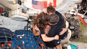 Tres astronautas, dos hombres y una mujer, se abrazan sonrientes en el interior de una nave espacial, con banderas de EE. UU. y Canadá visibles al fondo