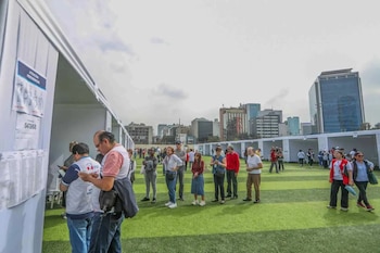 Multitud de personas de pie sobre césped artificial, esperando cerca de cabinas blancas de votación. Edificios de la ciudad y cielo gris de fondo