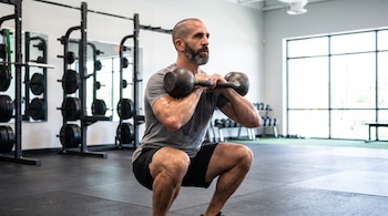 Hombre calvo con barba hace sentadilla con dos pesas rusas en un gimnasio, con estantes de pesas y ventanas grandes al fondo.