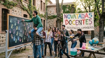 Estudiantes reunidos junto a una pizarra con huellas de manos, una pancarta que dice Novatadas Docentes y una mesa con globos terráqueos.
