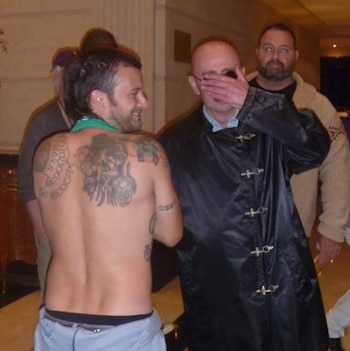 Hombre de espaldas sin camiseta mostrando tatuajes, hombre cubriendo su rostro, y hombre barbudo al fondo