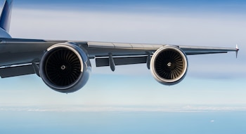 Un avión con un ala gris y dos motores grandes, de los que se ven las turbinas, vuela sobre un mar de nubes blancas y un cielo azul.