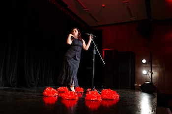Una mujer con cabello rojo viste ropa negra de pie junto a un micrófono en un escenario con grandes flores rojas en el suelo