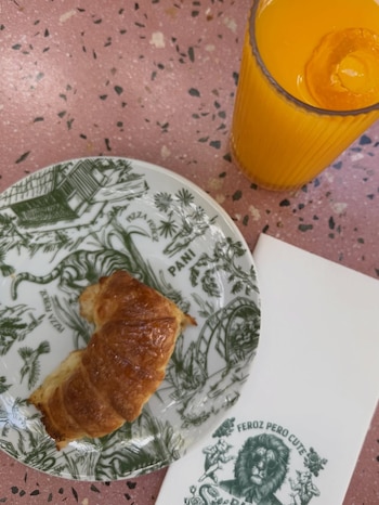 Vista cenital de un desayuno con un croissant en plato decorado, un vaso de zumo de naranja y una servilleta con logo de león sobre una mesa rosa