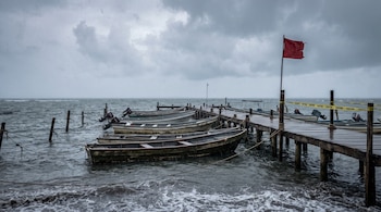 Muelle de madera con varias lanchas amarradas, mar agitado bajo lluvia y cielo cubierto, bandera roja en un poste y cinta amarilla de precaución.