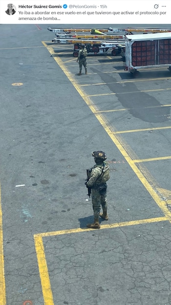 Vista aérea de dos militares en uniforme de camuflaje en una pista de aeropuerto, uno de espaldas con rifle y otro en la distancia, cerca de carros de equipaje