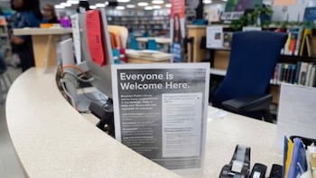 Primer plano de un mostrador de biblioteca con un letrero que dice 'Everyone is Welcome Here'. Al fondo, estanterías llenas de libros y personas borrosas