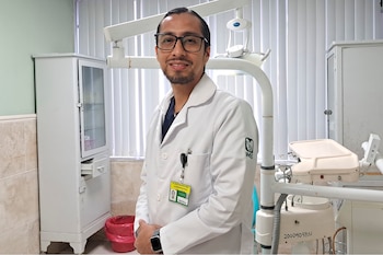 Hombre con bata blanca y gafas sonríe en una clínica. Detrás hay un gabinete blanco, persianas verticales y equipo médico