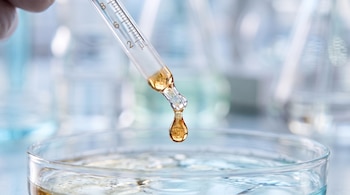 Mano con guante blanco sostiene una pipeta graduada, de la cual cae una gota de líquido ámbar en una placa de Petri con líquido transparente. Fondo de laboratorio borroso.