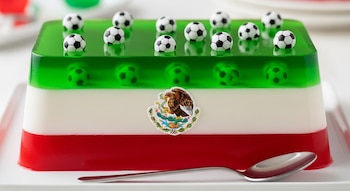 Gelatina rectangular con capas verde, blanca y roja, representando la bandera de México, adornada con balones de fútbol y el escudo nacional, con una cuchara al frente.