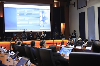 Personas sentadas en un auditorio, enfocadas en una pantalla grande que exhibe un sistema de software con formularios. Camarógrafos presentes al fondo