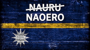 Una bandera de Nauru con fondo azul y banda horizontal amarilla. Contiene una estrella de doce puntas blanca y los textos "NAURU" tachado y "NAOERO".