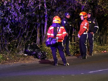 Dos bomberos con uniforme rojo y cascos y dos policías permanecen de pie de noche junto a una bolsa plástica negra en la carretera