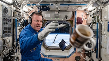 Astronauta con auriculares y guantes en un traje azul, flotando en una estación espacial. Una llave inglesa, una tableta y una taza de café flotan lejos. La Tierra se ve por la ventana.