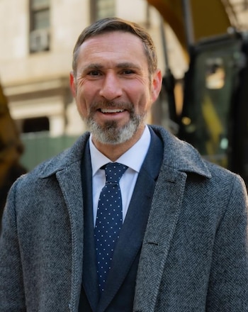 Primer plano de Jamie Torres-Springer, un hombre sonriente con barba, traje oscuro, corbata azul con puntos blancos y abrigo gris, con maquinaria al fondo