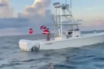 Una embarcación blanca con dos motores fuera de borda en el mar. Un hombre está a bordo, y hay dos banderas de buceo. Varias personas nadan cerca del barco