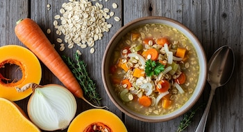 Toma cenital de sopa de avena y verduras en un bol. Alrededor: avena, zanahoria, calabaza, cebolla, hierbas y cuchara sobre madera rústica.