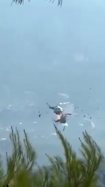 Dos helicópteros en el aire, uno de ellos en llamas y ambos con escombros, frente a un cielo brumoso y plantas verdes borrosas en primer plano