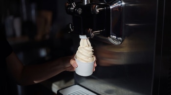 Una mano sostiene un vaso blanco de papel debajo del dispensador de una máquina metálica que sirve helado suave.