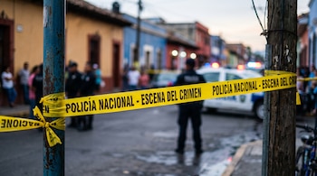 Cinta de seguridad amarilla con el texto Policía Nacional - Escena del crimen, atada a un poste frente a casas y una patrulla.