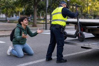 Un agente con chaleco reflectante coloca un monopatín en una plataforma junto a un joven de rodillas en la calle.
