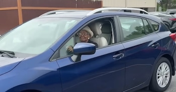 Susan Young Browne, una mujer afroamericana mayor con sombrero claro y gafas, sonríe desde el asiento del conductor de un coche azul oscuro