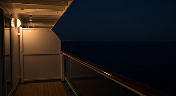 Vista desde el balcón de un camarote de crucero por la noche. Se ve la barandilla y parte de la estructura iluminada con el océano oscuro al fondo.
