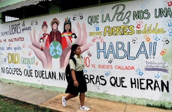 Uma jovem com mochila escolar e uniforme caminha da esquerda para a direita em frente a uma pintura branca com grafites coloridos sobre a paz e os direitos das crianças
