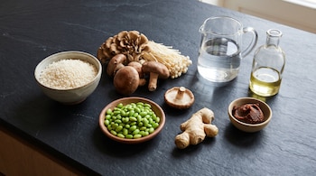 Cuencos con arroz, edamame y miso junto a setas variadas, jengibre, una jarra de agua y una botella de aceite sobre una mesa oscura.