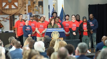Kathy Hochul habla tras un podio con el sello de Nueva York frente a un público y junto a personas con camisetas rojas y banderas