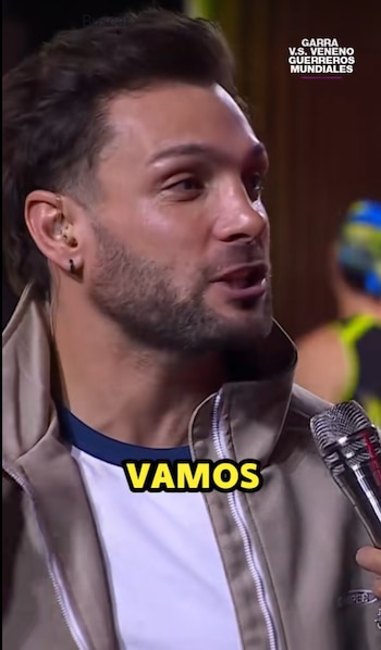 Primer plano de un hombre con barba corta, Nicola Porcella, hablando en un micrófono plateado. Viste una chaqueta beige y una camiseta con cuello azul