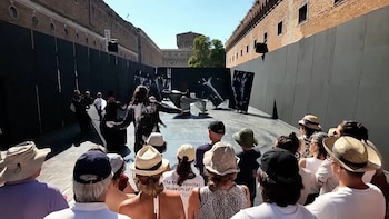 Escena teatral al aire libre con artistas en un escenario oscuro frente a una audiencia. Edificios de ladrillo y una torre en el fondo bajo cielo azul