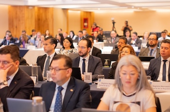 Una sala de conferencias con múltiples personas sentadas en mesas, usando laptops. Hombres y mujeres en atuendo formal. Botellas de agua en las mesas