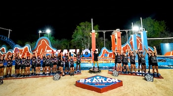 Participantes con ropa deportiva y los brazos levantados rodean al conductor en un set nocturno sobre arena con la palabra Exatlón México.