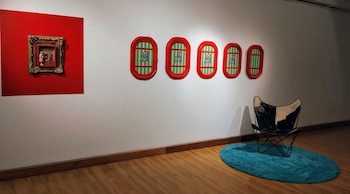 Vista de una sala de exposición con una gran obra de arte en rojo, cuatro marcos ovalados con dibujos, una silla y una alfombra azul en un piso de madera