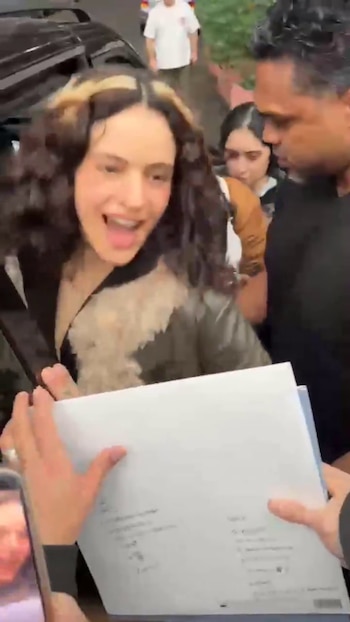 Una mujer joven con cabello oscuro y mechas claras, Rosalía, sonríe ampliamente mientras personas a su alrededor sostienen papeles y un teléfono