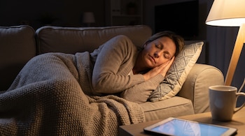 Mujer durmiendo en un sofá, cubierta con manta, cabeza sobre cojín. Lámpara de pie, taza y tablet con pantalla encendida sobre mesa.