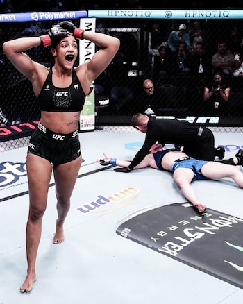 Una luchadora de UFC celebra con los brazos en alto y la boca abierta, mientras su oponente yace noqueada en el suelo del octágono y un árbitro la asiste