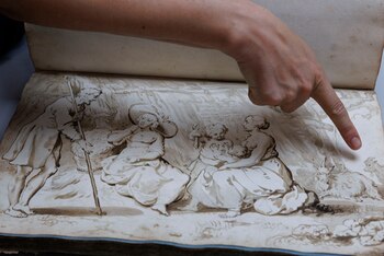Una mano de piel clara señala un dibujo antiguo de estilo sepia en un libro abierto que muestra un hombre, dos mujeres, dos niños y dos cabras