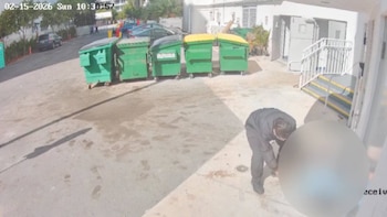 Cámara de seguridad muestra a un hombre agachado frente a contenedores de basura verdes en un patio de servicio, con un edificio y calle al fondo